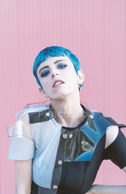 Young woman in trendy alternative dress touching short blue hair and looking at camera while standing against pink wall on city street — Stock Photo