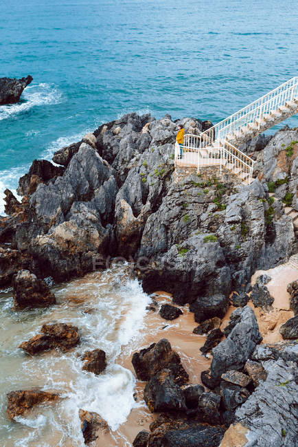 Side view of woman admiring waving and crashing blue sea water in bay while standing on stairway on cliff — Stock Photo
