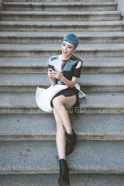 Desde arriba de mujer joven con el pelo azul corto y en vestido futurista de moda con teléfono en las escaleras de la calle - foto de stock