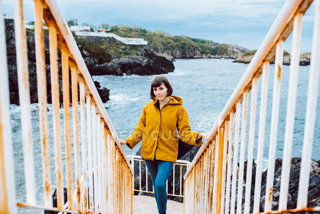 Young woman in yellow jacket standing on rusty stairway against bay with waving sea water — Stock Photo
