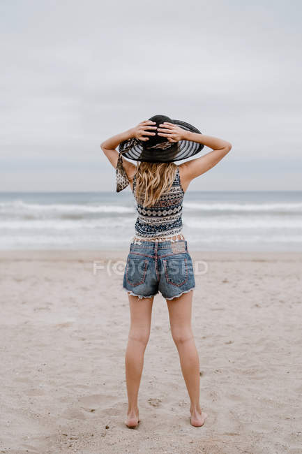 Rückseite der attraktiven Frau mit Oberteil und kurzen Hosen, die mit schwarzem Hut am Sandstrand steht — Stockfoto