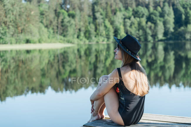 Боковой вид счастливой молодой женщины в черном купальнике и шляпе, сидящей на деревянном пирсе и любующейся видом на озеро на ясном голубом небе и лесном фоне — стоковое фото