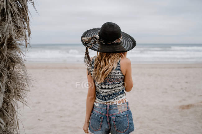 Вид сзади на привлекательную женщину в топе и шортах, стоящую на песчаном берегу моря в черной шляпе — стоковое фото