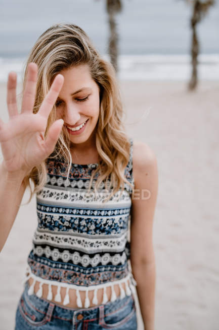Cheerful blonde woman in colorful top and jean shorts blocking her face with hand on seashore — Stock Photo