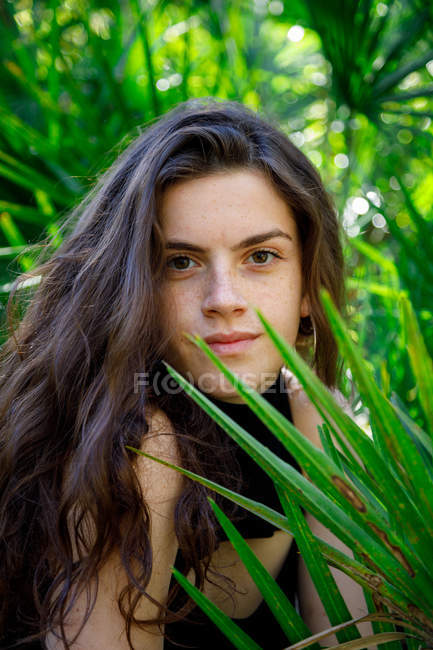 Ritratto di bruna sorridente giovane donna seduta in cespugli verdi tropicali — Foto stock