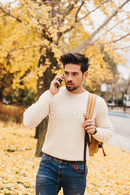 Handsome young man with trendy hairstyle walking on autumn leaves and speaking on smartphone on daytime — Stock Photo