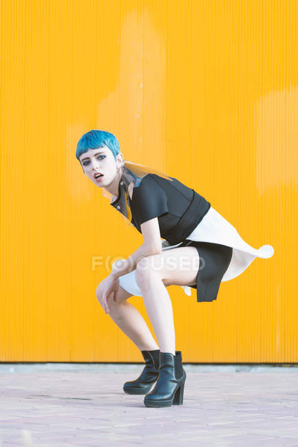 Sinnliche junge Frau im trendigen futuristischen Kleid, die in die Kamera blickt, während sie auf Knien auf dem Bürgersteig vor einer knallgelben Wand steht — Stockfoto