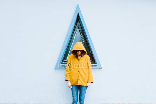 Mujer joven en abrigo cálido amarillo sonriendo y mirando hacia abajo mientras está de pie contra la ventana del triángulo y la pared gris del edificio - foto de stock