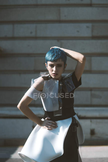 Young woman in futuristic dress touching head and short blue hair while standing near building on sunny day on city street — Stock Photo