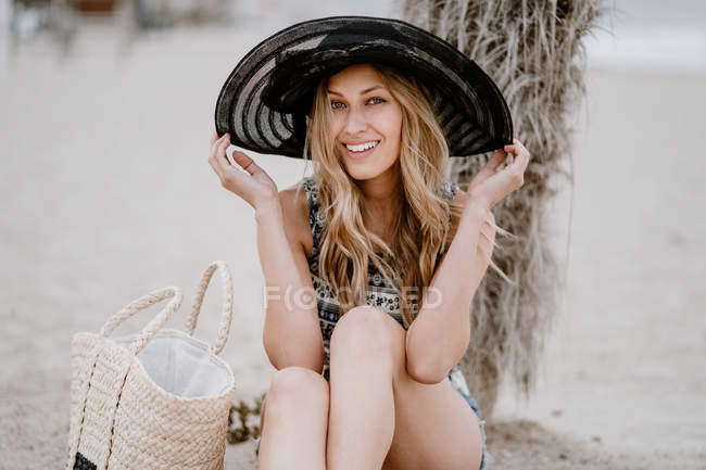Блондинка в черной шляпе сидит на песке с летней сумкой и смотрит в камеру — стоковое фото