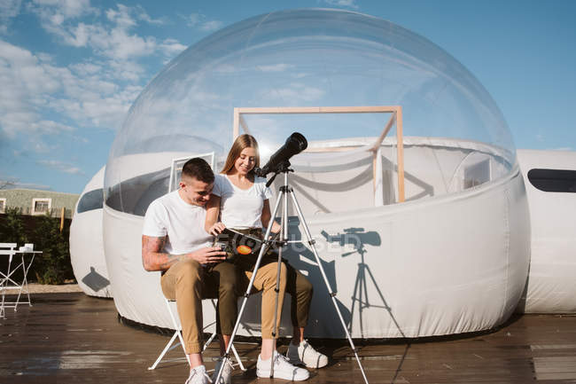 Стильная женщина, сидящая на коленях бойфренда и читающая на телескопе в небе возле отеля 