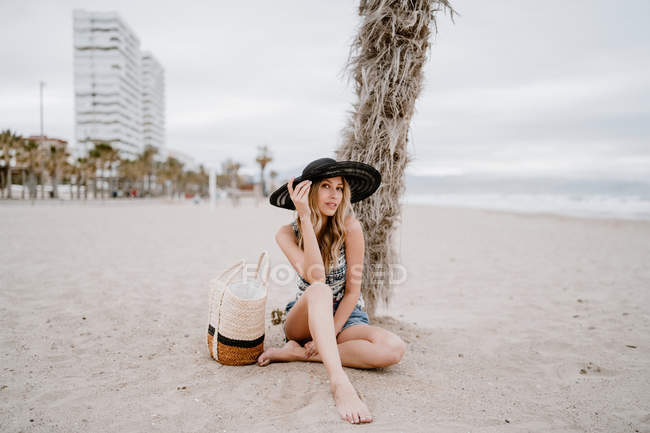Mujer rubia en sombrero negro sentado en la arena con bolsa de verano y mirando a la cámara - foto de stock