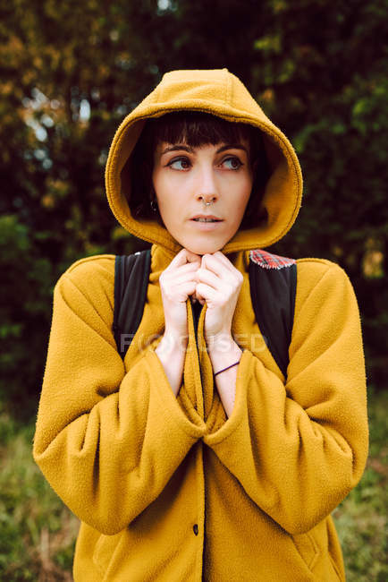 Frau hüllt sich in gelben Kapuzenmantel und schaut weg, während sie an kalten Tagen vor verschwommenem Hintergrund der Natur steht — Stockfoto