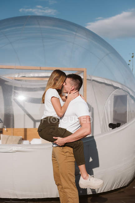 Vista lateral de un hombre apasionado sosteniendo y abrazando a una mujer joven mientras está de pie frente al glamping transparente romántico - foto de stock