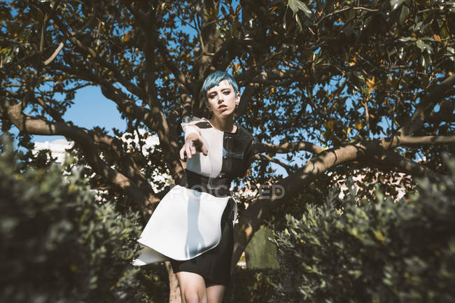 Desde abajo mujer joven en vestido futurista mirando a la cámara mientras está de pie cerca de los árboles en desenfoque - foto de stock