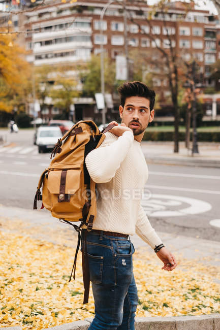 Модний чоловік в джинсах і білому светрі тримає помаранчевий рюкзак на плечі і дивиться в осіннє місто — стокове фото