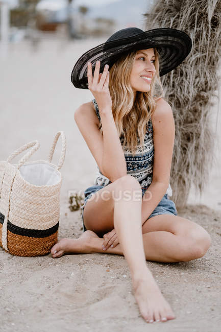 Mujer rubia en sombrero negro sentado en la arena con bolsa de verano y mirando hacia otro lado - foto de stock
