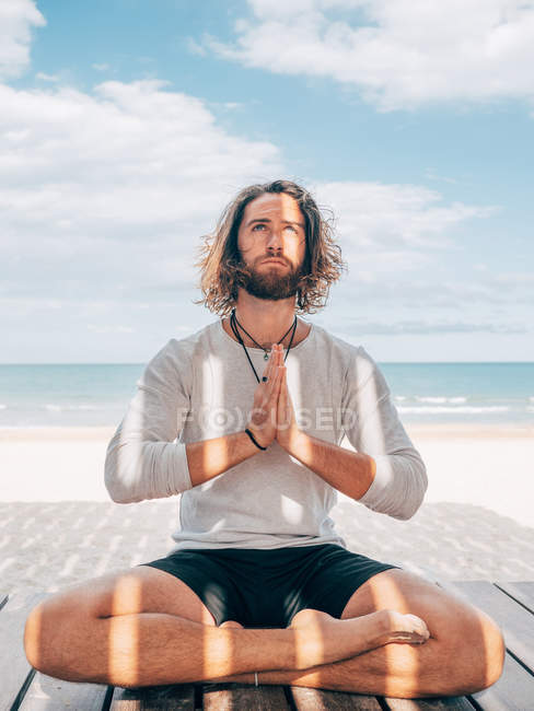 Adult bearded man meditating while sitting in lotus pose on wooden pier by seashore with legs crossed and looking up — Stock Photo