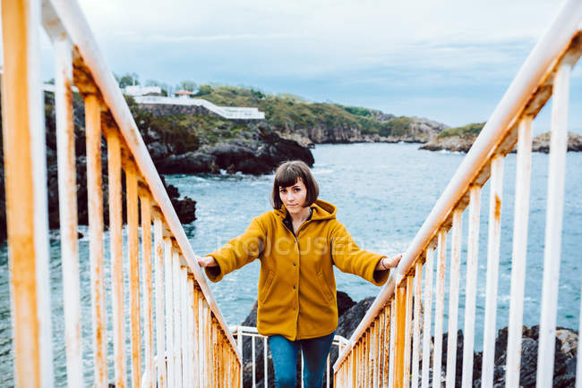 Jovem mulher de casaco amarelo em pé na escada enferrujada contra a baía com água do mar ondulando — Fotografia de Stock