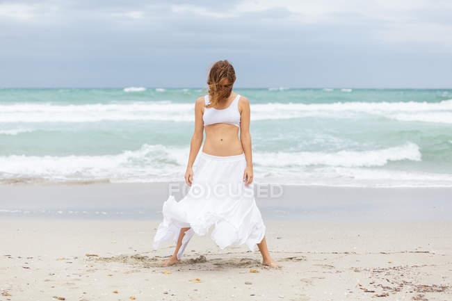 Привлекательная женщина в белом наряде танцует на песке возле моря — стоковое фото