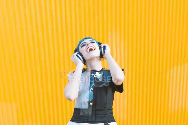 Fröhliche junge Frau im trendigen alternativen Kleid lächelt und hört Musik mit Kopfhörern gegen die gelbe Wand — Stockfoto