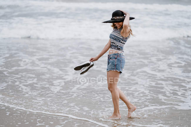 Mulher atraente em chapéu preto segurando sapatos enquanto desfruta de uma vista pitoresca do oceano olhando para baixo — Fotografia de Stock