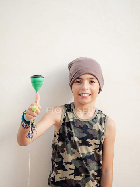 Smart stylish boy in hat easily spinning green top while looking at camera on white background — Stock Photo