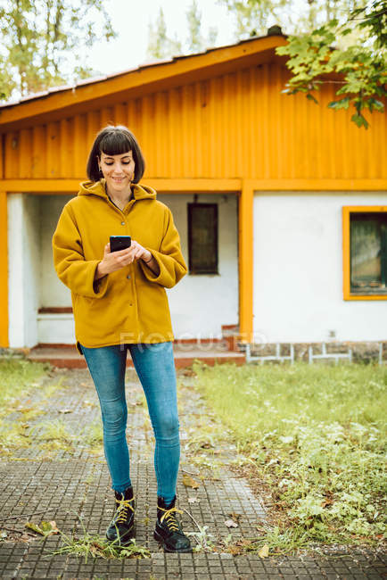 Mujer joven en traje casual sonriendo y navegando teléfono inteligente, mientras que de pie en el camino de baldosas exterior encantadora cabaña - foto de stock