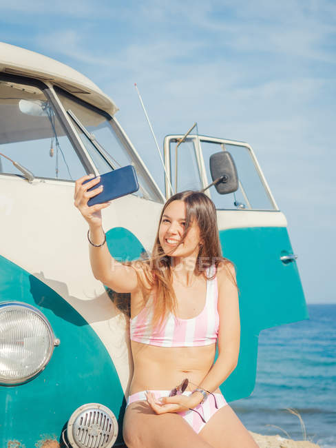 Mulher bronzeada encantadora atraente levando selfie e sorrindo perto de carro na praia de areia em dia brilhante — Fotografia de Stock