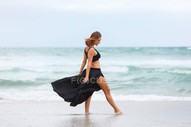 Элегантная женщина в черном наряде танцует на песке у моря — стоковое фото