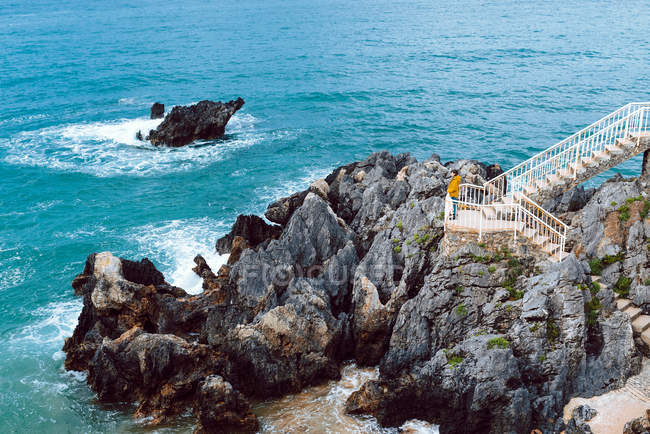 Vista lateral de la mujer admirando agitar y estrellarse el agua azul del mar en la bahía mientras está de pie en la escalera en el acantilado - foto de stock