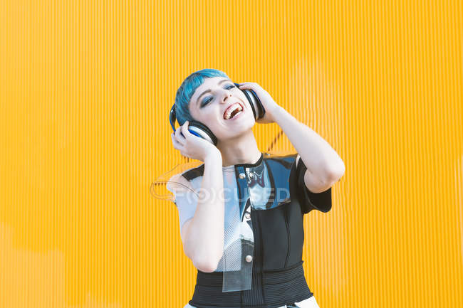 Cheerful young woman in trendy alternative dress smiling and listening to music in headphones against yellow wall — Stock Photo