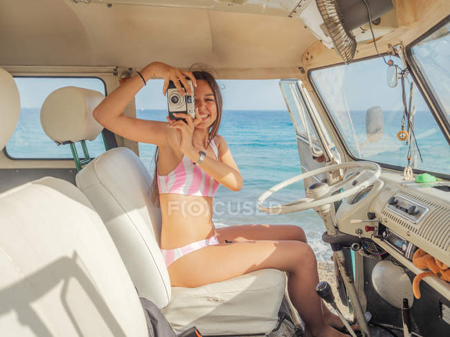 Frau im Badeanzug mit Kamera lächelt und macht Foto im weißen Vordersitz des Autos am Meer bei sonnigem Tag — Stockfoto