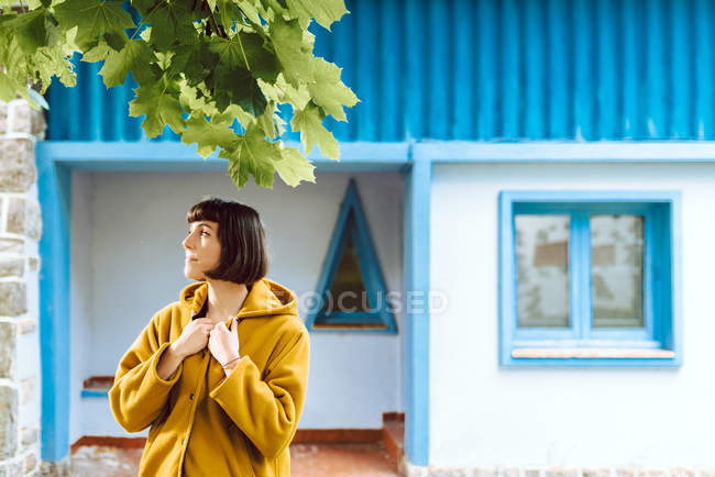 Junge Frau in gelbem warmen Mantel lächelt und schaut weg, während sie vor grauer Wand steht — Stockfoto