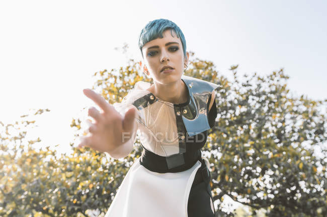 De baixo jovem mulher com cabelo azul curto e em vestido futurista dobrando para a frente e tentando tocar câmera no fundo borrado — Fotografia de Stock