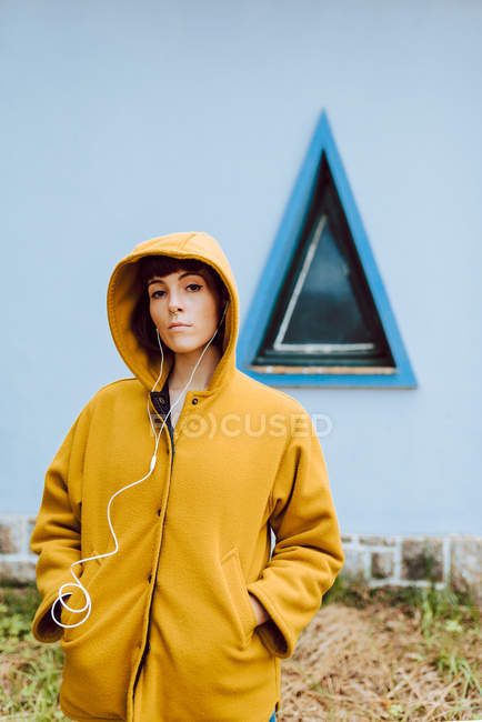 Jovem mulher de casaco quente amarelo ouvindo a música e olhando para a câmera enquanto está de pé contra a janela do triângulo e parede cinza — Fotografia de Stock