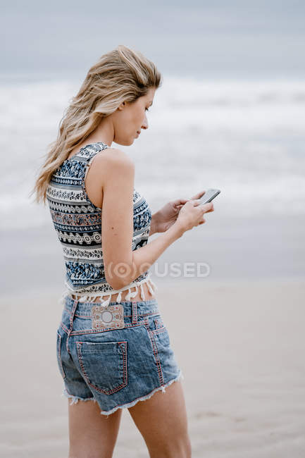 Back view of young blonde cheerful woman standing and using smartphone on sea background — Stock Photo