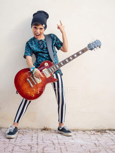 Cheeky chico excitado activo en ropa colorida tocando la guitarra mostrando dos dedos en el fondo de la pared blanca - foto de stock