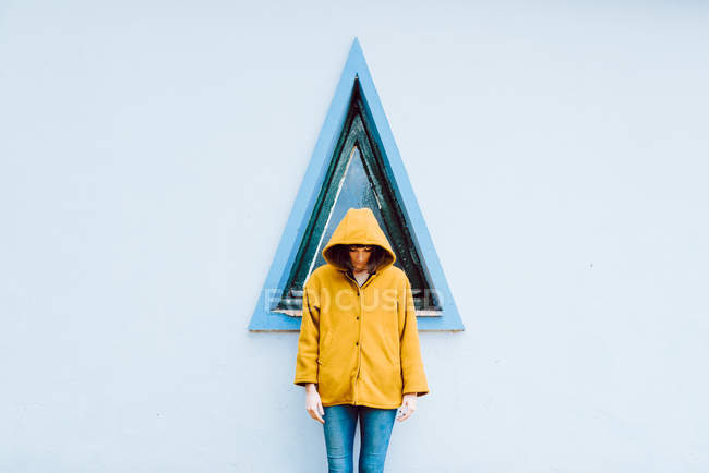 Young woman in yellow warm coat smiling and looking down while standing against triangle window and gray wall of building — Stock Photo
