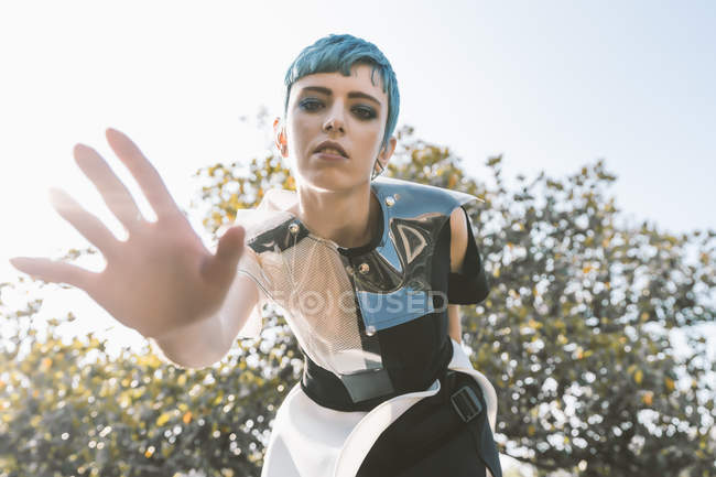 From below young woman with short blue hair and in futuristic dress bending forward and trying to touch camera on blurred background — Stock Photo