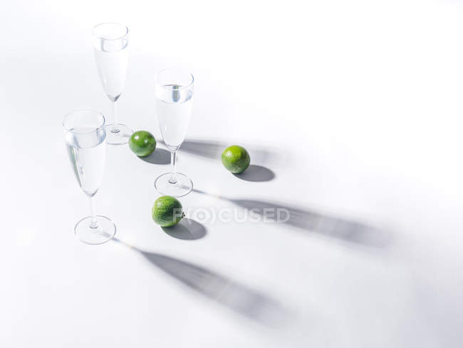 Kontrastierende Schatten aus Wassergläsern und grünen reifen Limetten auf weißem Hintergrund — Stockfoto
