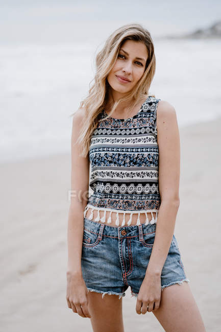 Весела блондинка в барвистих топах і джинсових шортах позує під час відпочинку на морському узбережжі — стокове фото