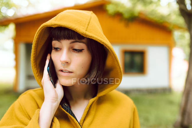 Mujer joven en traje casual sonriendo y hablando por teléfono inteligente, mientras que de pie en el camino de baldosas fuera encantadora casa de campo en el día de otoño en el campo - foto de stock