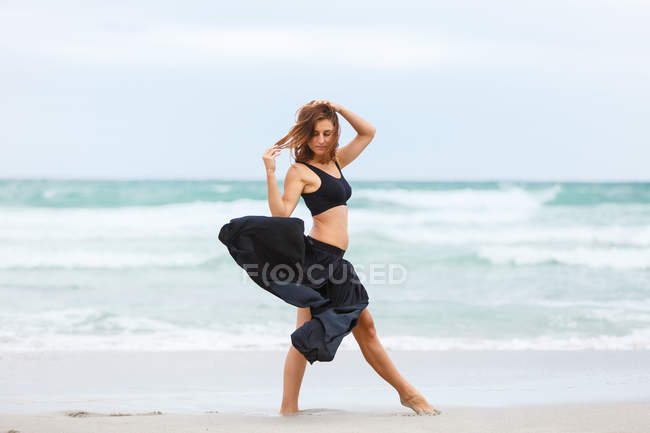 Elegante donna in abito nero che danza sulla sabbia vicino al mare ondulante — Foto stock