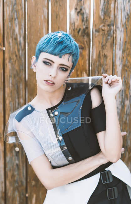 Young woman with short blue hair wearing futuristic dress and looking at camera while standing near shabby lumber fence — Stock Photo