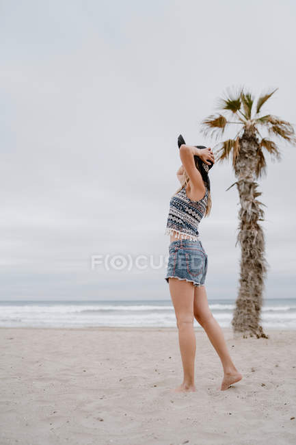 Mujer atractiva vistiendo top y pantalones cortos bailando en playa de arena con sombrero negro - foto de stock