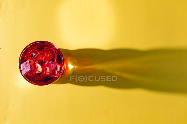 Do acima mencionado vidro com bebida vermelha saborosa e cubos de gelo na superfície amarela brilhante na luz — Fotografia de Stock