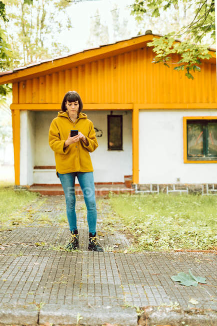 Mujer joven en traje casual sonriendo y navegando teléfono inteligente, mientras que de pie en el camino de baldosas fuera encantadora casa de campo en el día de otoño en el campo - foto de stock