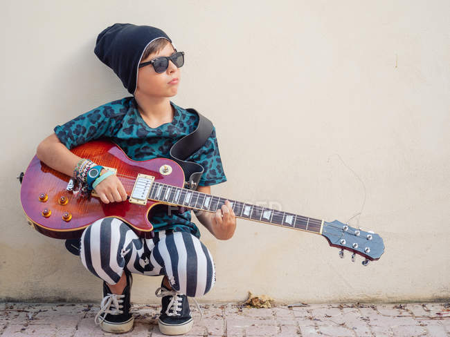 Cheeky active excited boy in colorful clothes playing guitar, leaning on background of white wall — Stock Photo