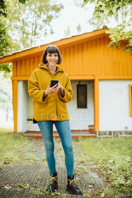 Молода жінка в повсякденному вбранні посміхається і переглядає смартфон, стоячи на плитці біля прекрасного котеджу в осінній день в сільській місцевості — стокове фото